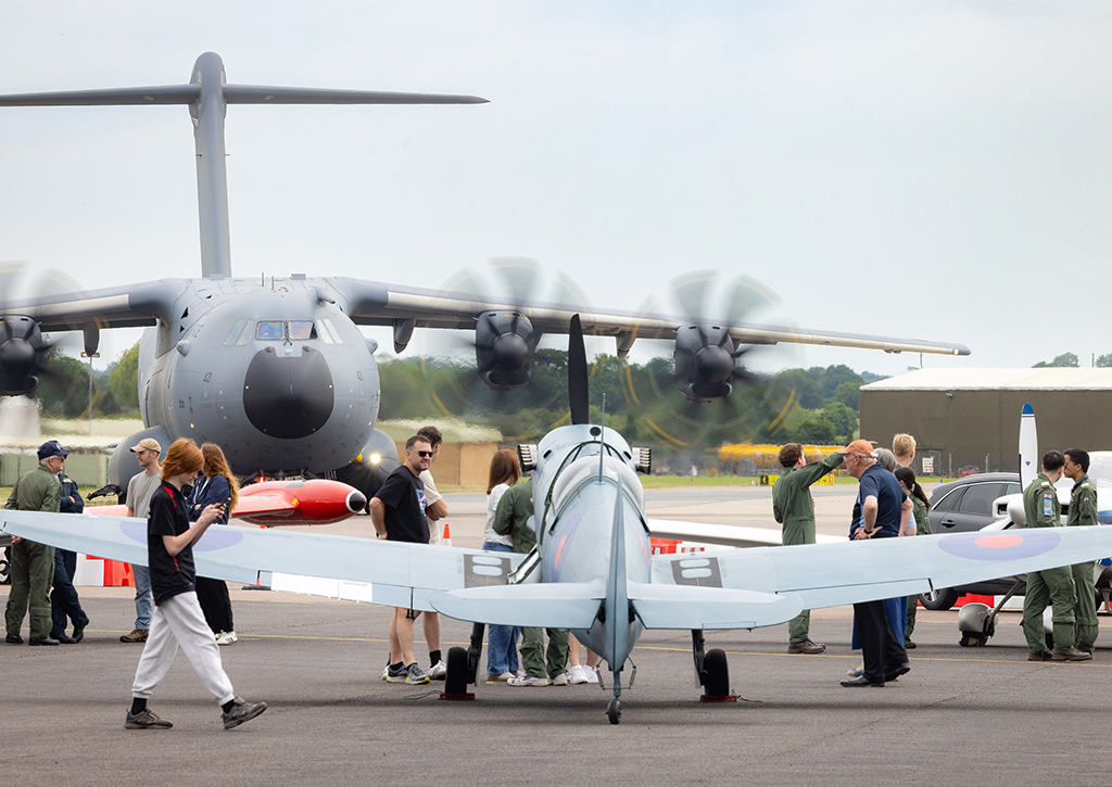 A rear view of a light blue WWII Spitfire fighter aircraft parked on runway tarmac, surrounded by people, with with a massive RAF Atlas A400M military transport aircraft propellers turning in the background.